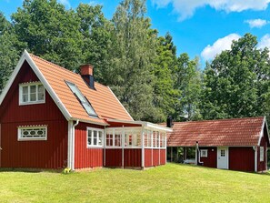 Ein rotes Bauernhaus mit geneigtem Dach und einem großen Fenster, umgeben von einer Wiese und Bäumen.