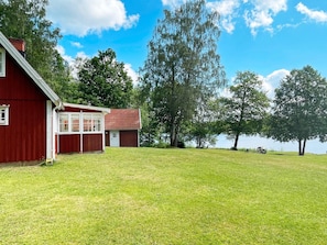 Ein rotes Bauernhaus mit weißer Tür und Fenstern, ein kleiner angebauter Anbau mit rotem Dach, und ein grasbewachsenes Feld mit Bäumen und einem Gewässer im Hintergrund.