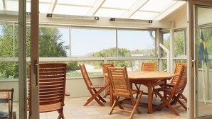 A sunroom with a large table and chairs, large windows, and a view of greenery and a road. - 4 Star Holiday Home in Hamburgsund (Hamburgsund)