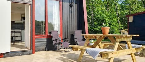 Una terraza de madera con una mesa de picnic y sillas, una pequeña planta en maceta, y un edificio con ventana de marco rojo y puerta que da a una zona de cocina.