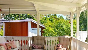 Eine überdachte Veranda mit Korbmöbeln, einem weißen Zaun und einem roten Gebäude im Hintergrund.