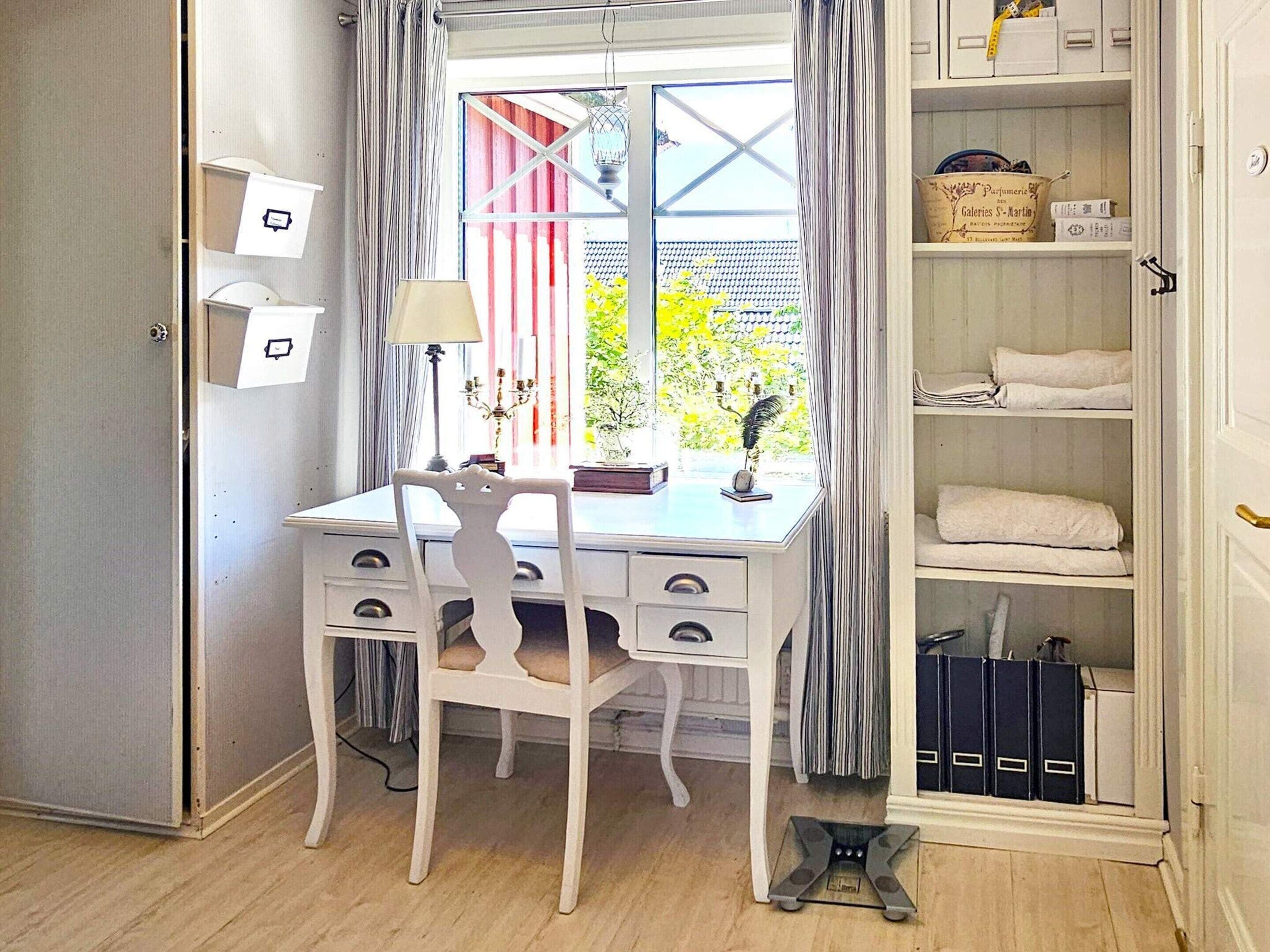 A room with a desk, chair, window with curtains, and a shelving unit with storage boxes.