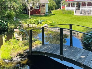 A wooden bridge over a pond with a garden and a house in the background. - 4 Star Holiday Home in Myggenas (Myggenäs)