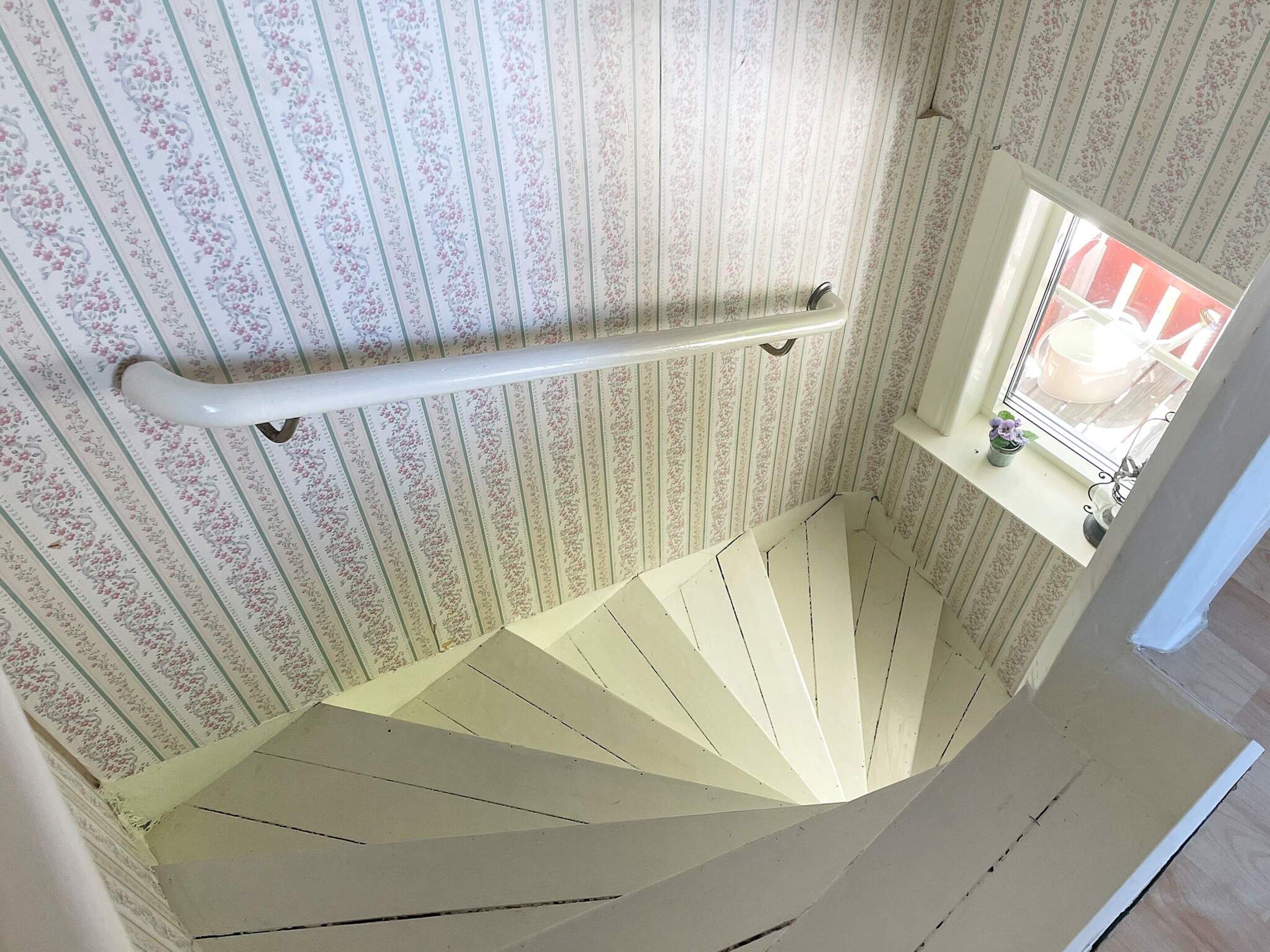 A spiral staircase with a handrail, a window with a view of the outside, and a floral wallpaper pattern.
