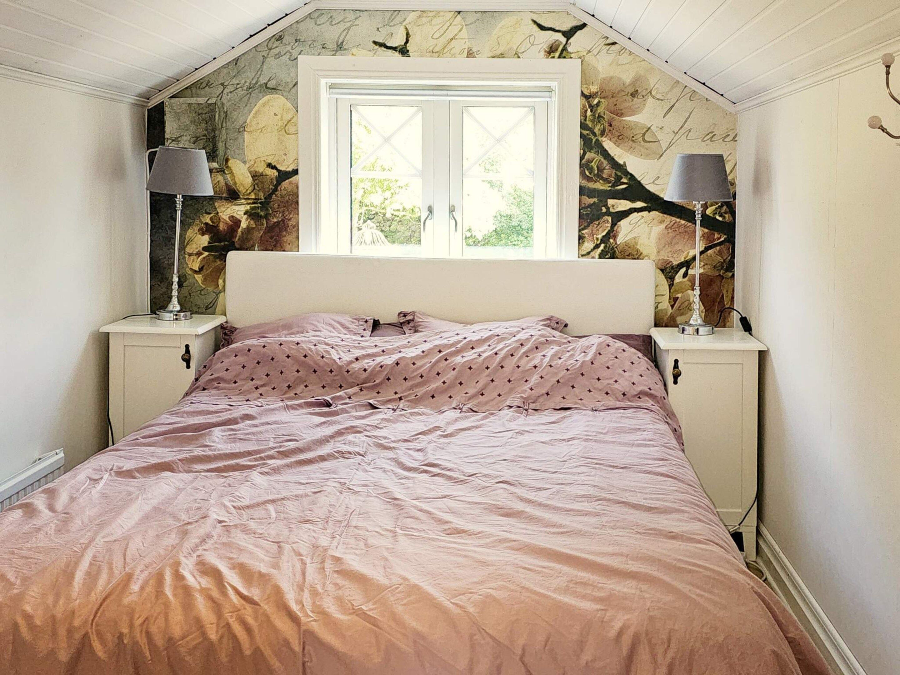 A bedroom with a bed, two bedside lamps, a window with a view, and a wall with a tree branch design.