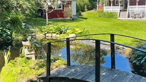 A wooden bridge over a pond with a statue, surrounded by lush greenery and a red building with a white porch. - 4 Star Holiday Home in Myggenas (Myggenäs)
