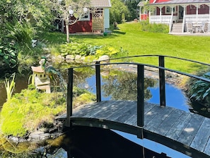 A wooden bridge over a pond with a statue, surrounded by lush greenery and a red building with a white porch. - 4 Star Holiday Home in Myggenas (Myggenäs)