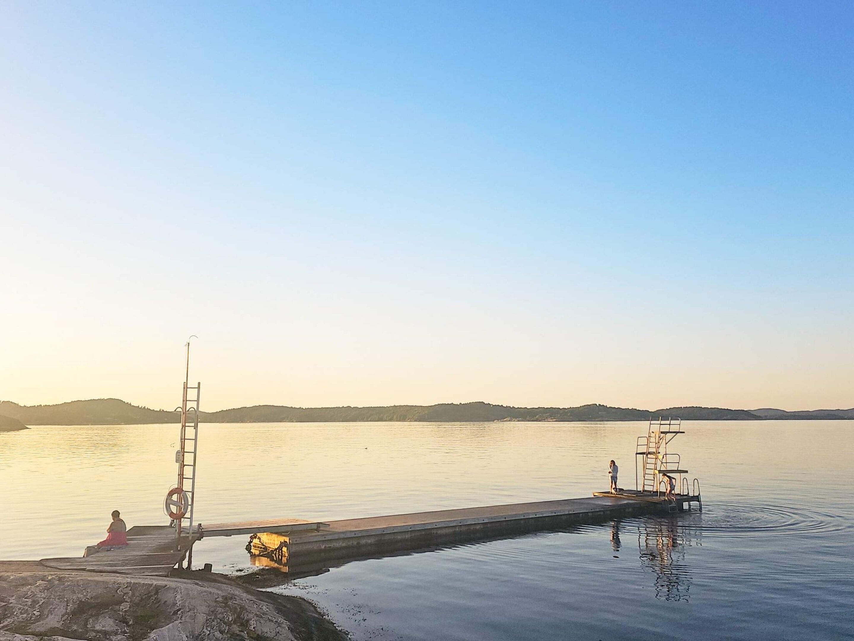 Eine Mole, die sich in ruhiges Wasser erstreckt, mit einer sitzenden Person am Rand und einer stehenden Person am Steg.