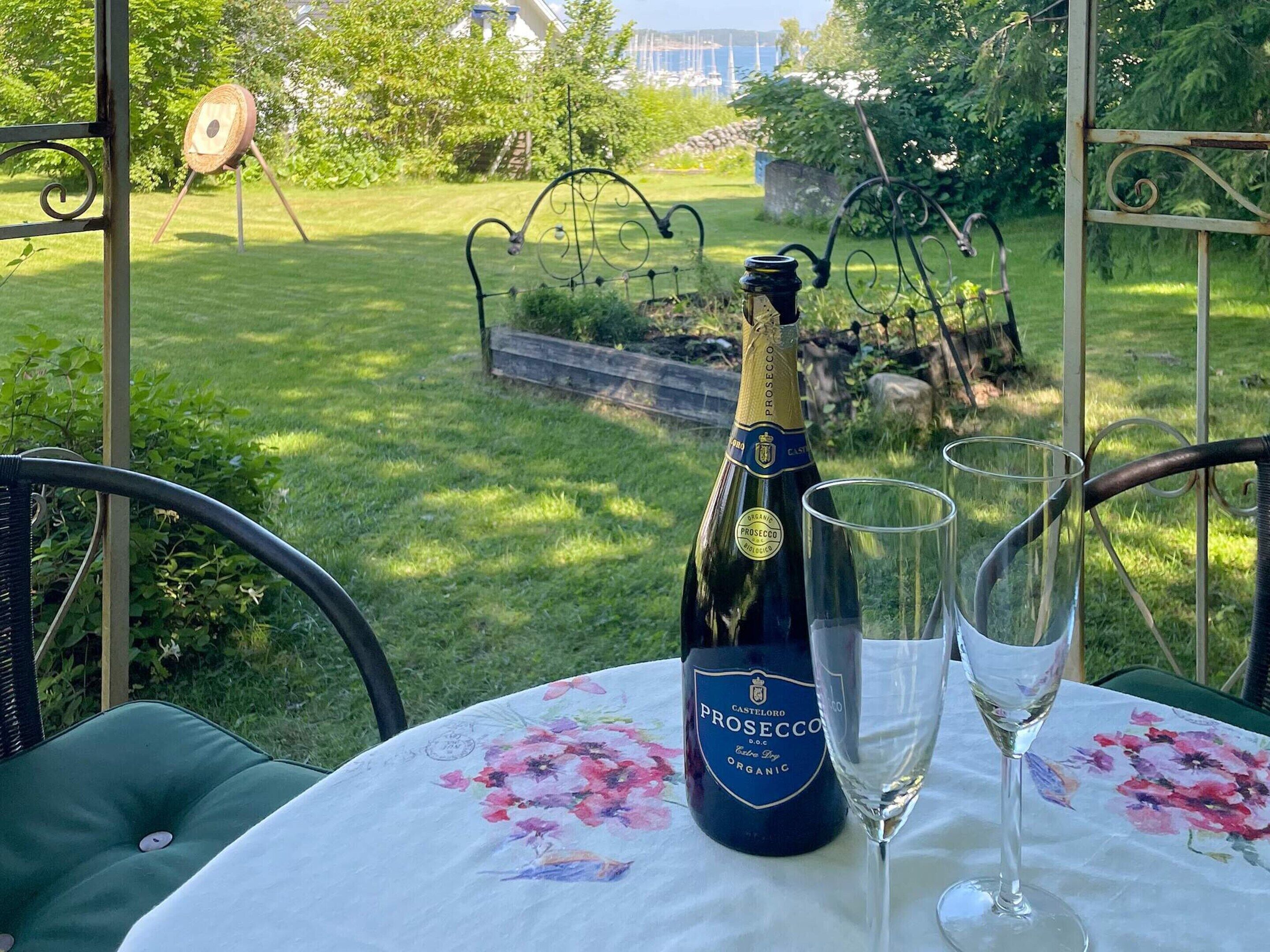 A bottle of Prosecco and two glasses on a table with a floral tablecloth, set outdoors with a garden view.