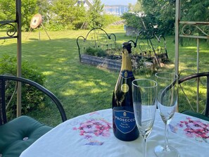 Une bouteille de Prosecco et deux verres sur une table recouverte d’une nappe à motifs floraux, le tout installé à l’extérieur avec vue sur un jardin.