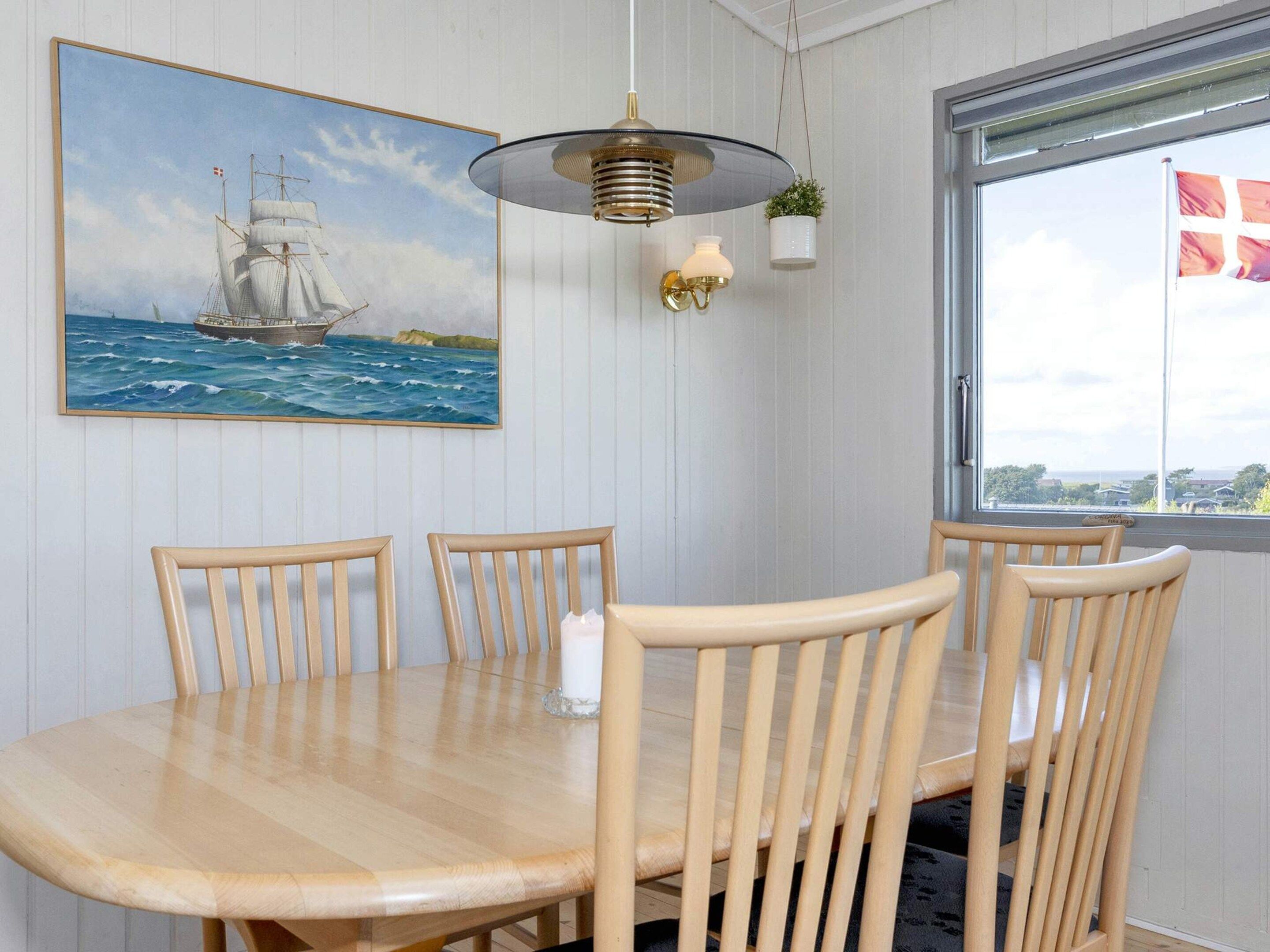A dining area with a wooden table and chairs, a painting of a sailing ship, a hanging light fixture, and a window with a view.