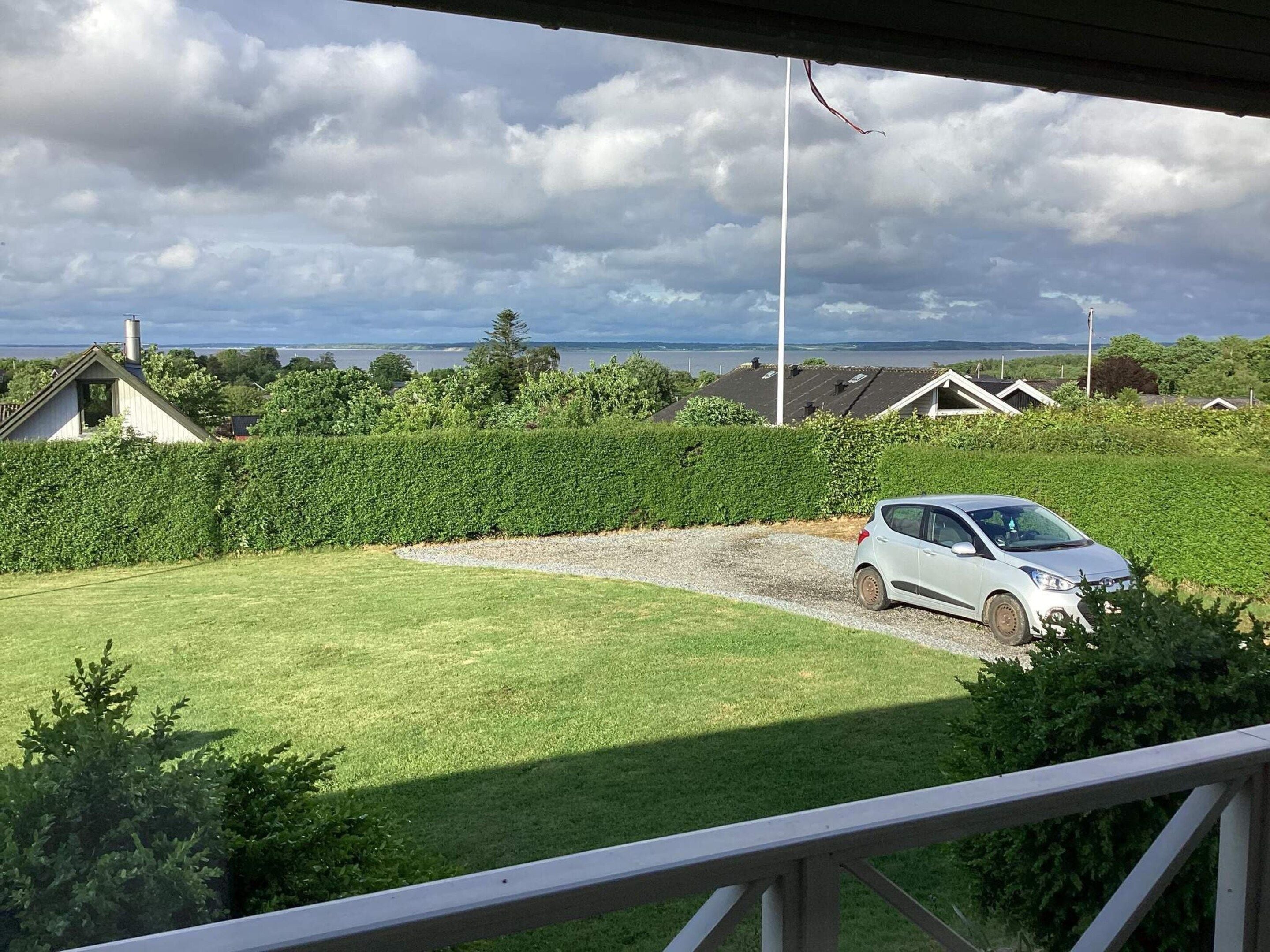 A car parked on a gravel driveway with a grassy lawn, a hedge, and a view of houses and a body of water in the distance.