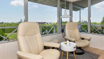 A room with two beige leather chairs, a small white table with a candle, and a view of greenery and a hammock outside.
