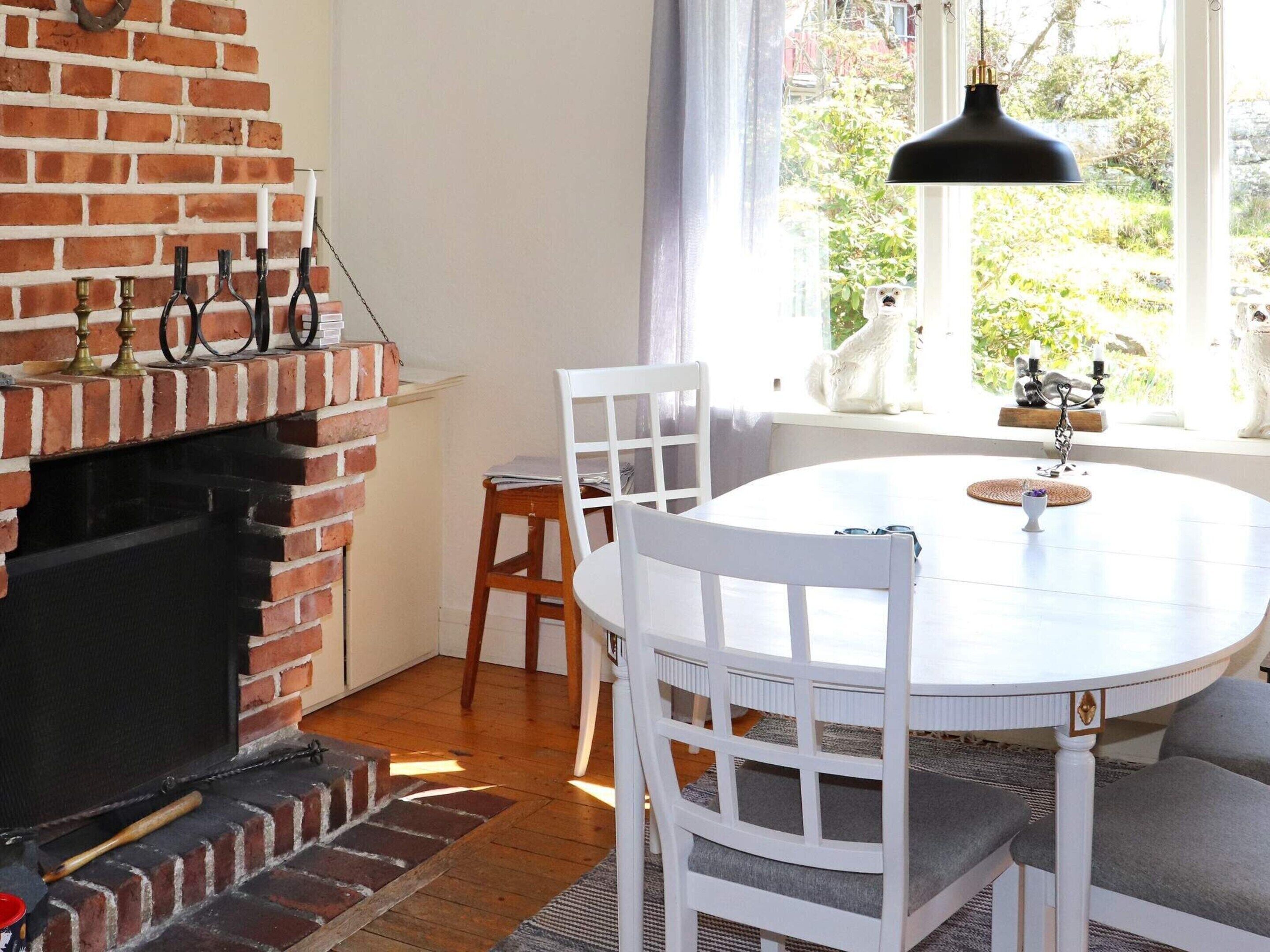 A cozy room with a brick fireplace, white chairs, a round table, and a view of greenery through a window.