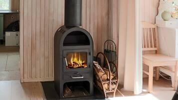 A wood-burning stove with a fire burning inside, a chair with a woven seat, and a wooden cabinet in the background.