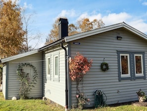 Une maison de plain-pied avec une cheminée, une fenêtre visible et un petit jardin agrémenté d’un tuyau d’arrosage et d’une plante en pot.