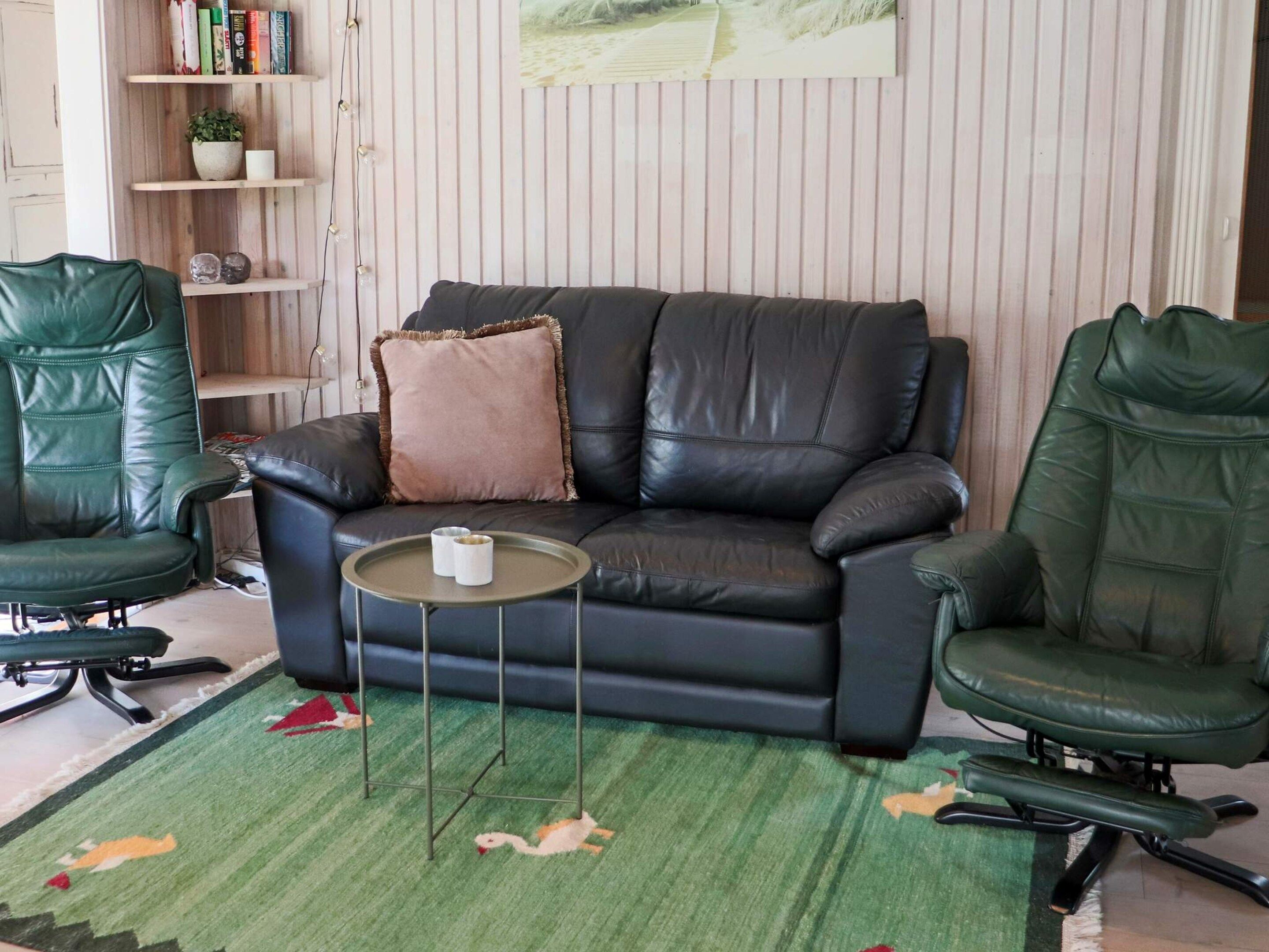 A living room with a black leather sofa, two green leather chairs, a small round table, a green rug with a swan design, and a bookshelf with books and decorative items.