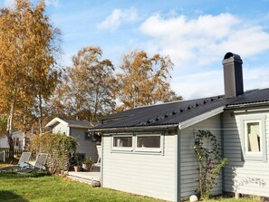 Ein eingeschossiges Haus mit dunklem Dach, Schornstein und einer kleinen Veranda mit zwei Stühlen.