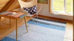 Une pièce avec un plafond en pente, une chaise dotée d’un coussin à motifs étoilés, une petite table surmontée d’un tapis coloré, un tableau accroché au mur et une fenêtre donnant sur un paysage verdoyant.