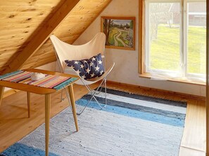 A room with a sloped ceiling, a chair with a star-patterned cushion, a small table with a colorful mat, a painting on the wall, and a window with a view of a green landscape. - 4 Person Holiday Home in Holmsjo (Holmsjö)