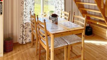 Una mesa de comedor de madera con sillas, un jarrón con flores y vista a un jardín a través de una ventana con cortinas estampadas.