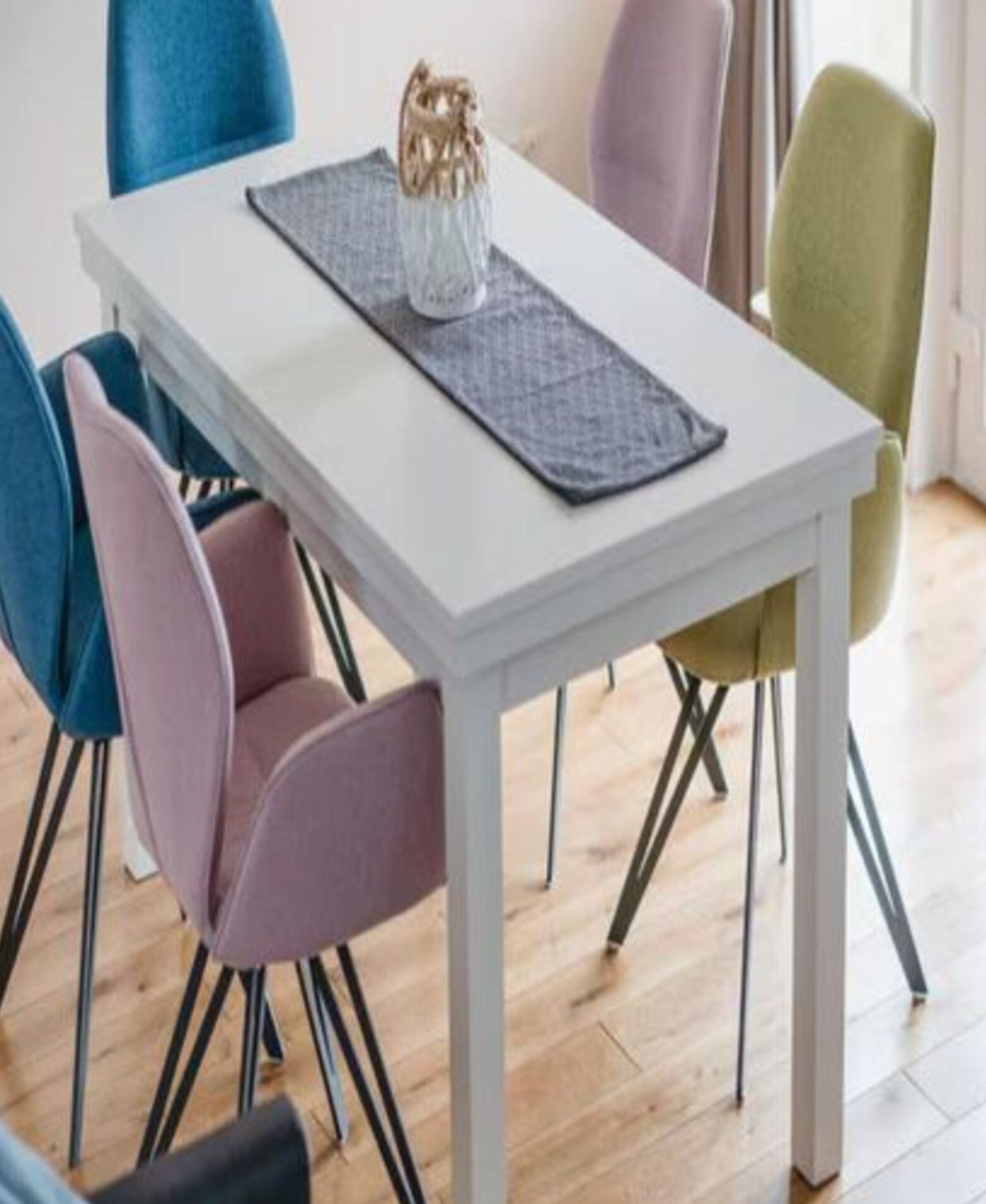 Ein Esstisch mit weißer Tischdecke, einer dekorativen Vase und einer Platzauflage. Darum herum sind vier Stühle in verschiedenen Farben platziert.
