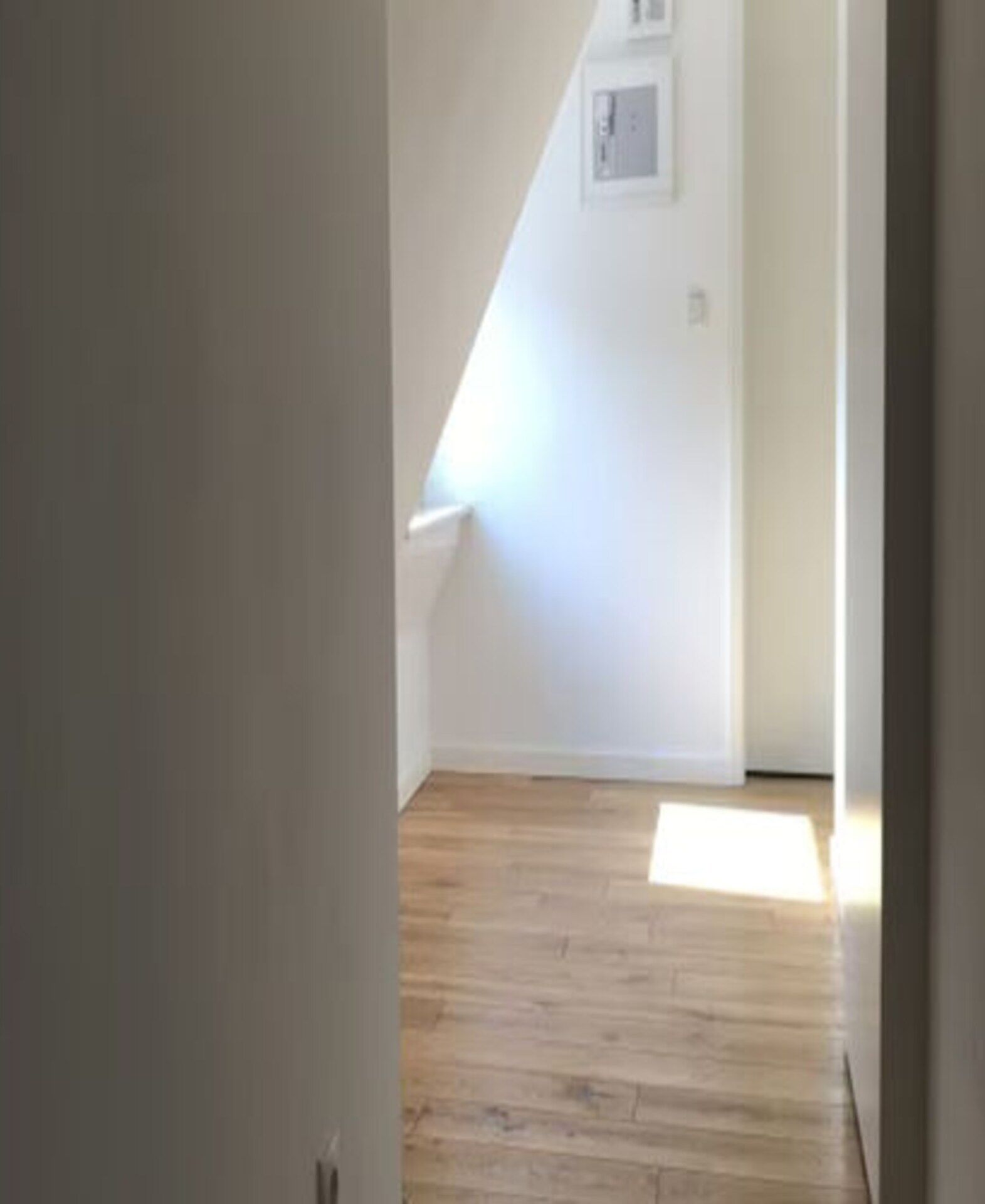 A narrow hallway with wooden flooring leading to a room with a sloped ceiling and a small window.