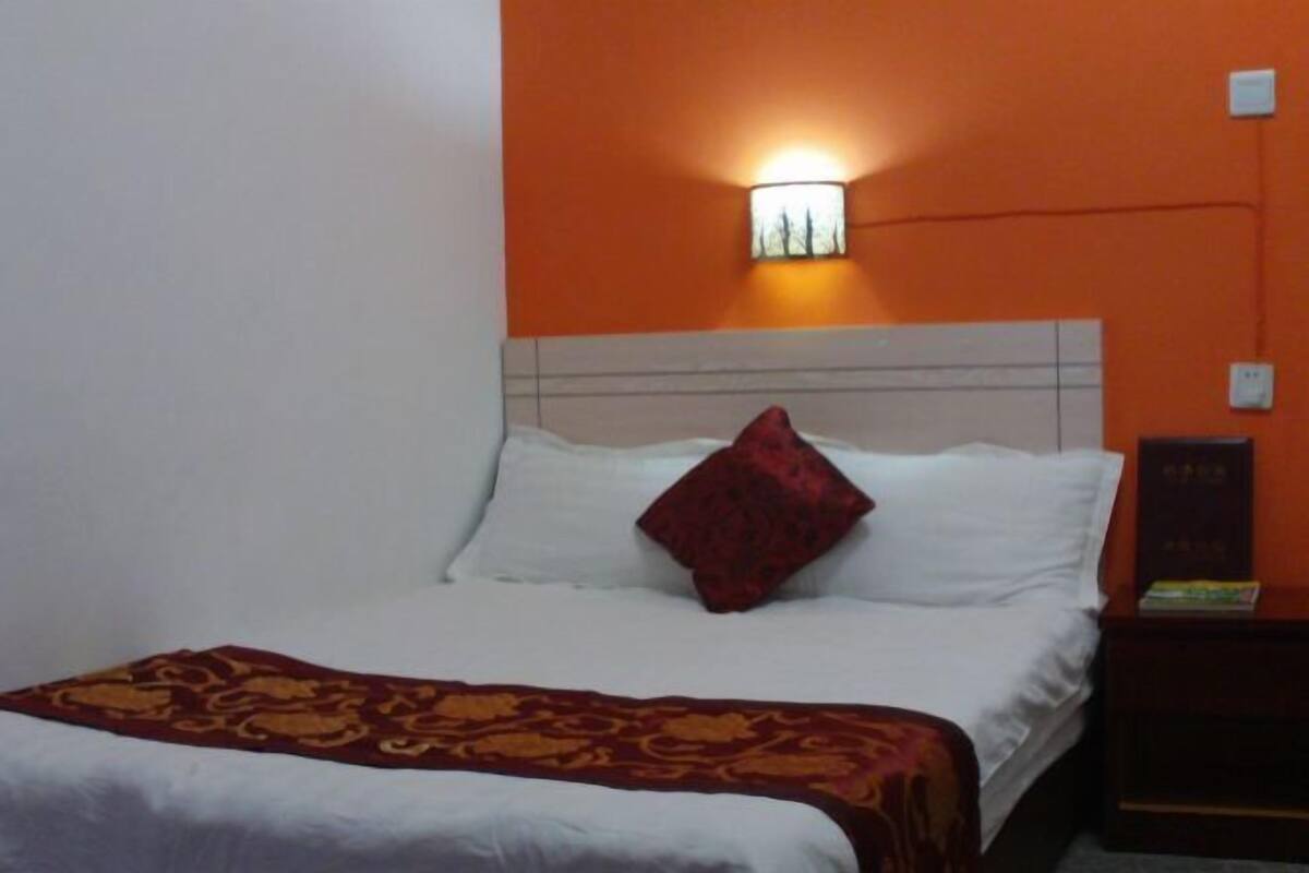 A hotel room with a bed, a red pillow, a patterned throw blanket, a nightstand with a book, and an orange wall with a wall-mounted light.