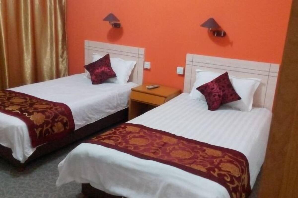 Two single beds with white linens and red patterned bedspreads, a wooden nightstand, and a wall-mounted lamp.