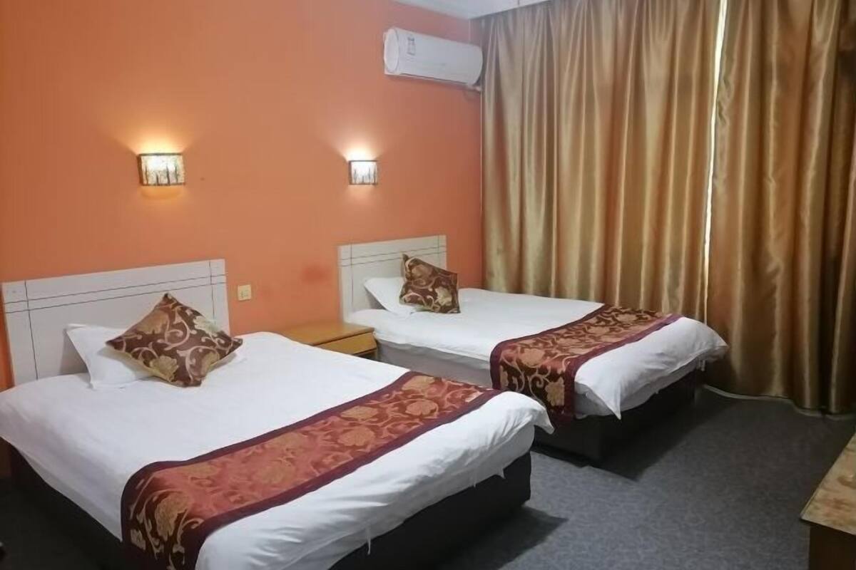A hotel room with two beds, each with a white mattress and a brown patterned bedspread. There are two wall-mounted light fixtures on the orange walls.