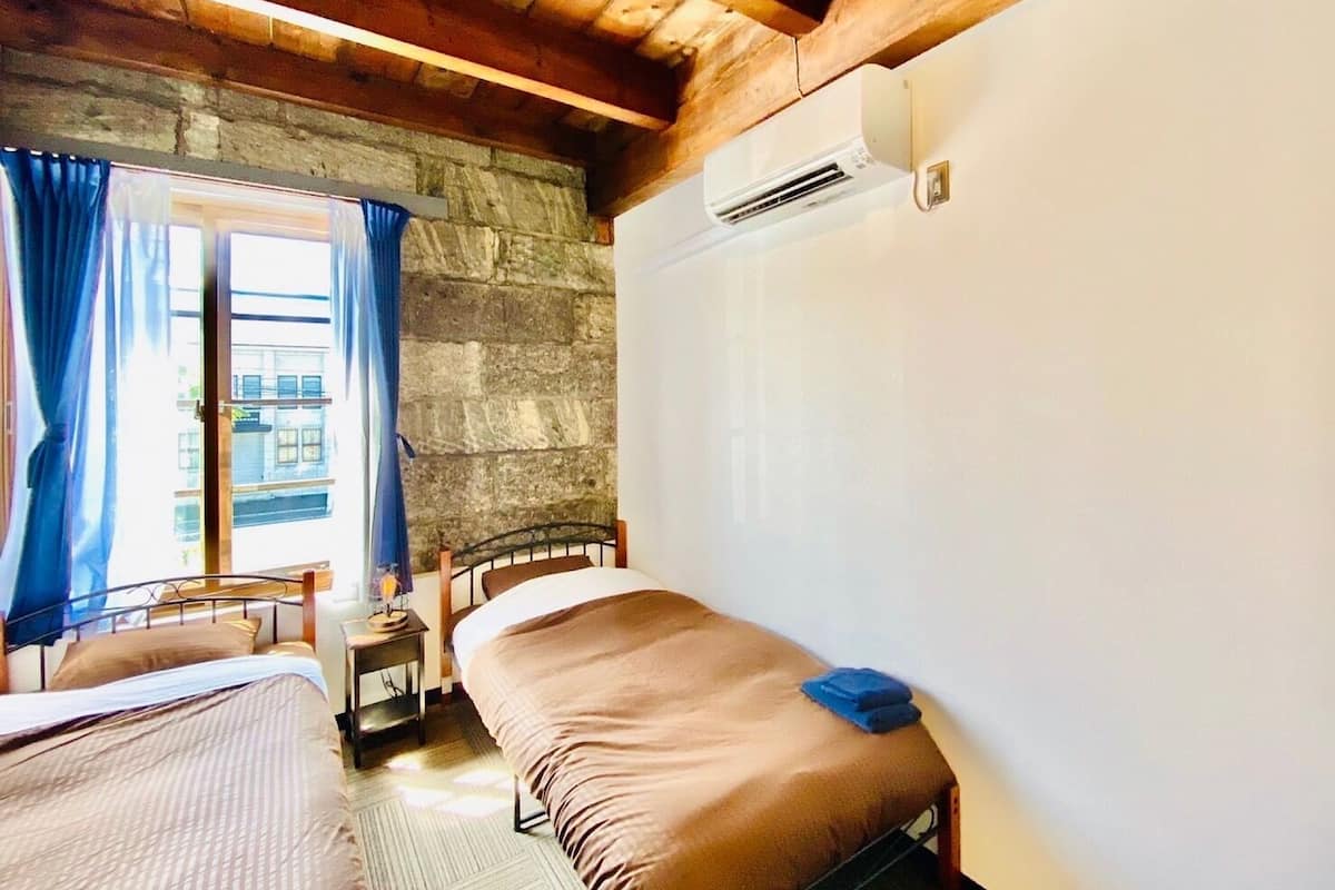 Une pièce avec un lit, une petite table avec une lampe, une fenêtre avec des rideaux et un mur en pierre.