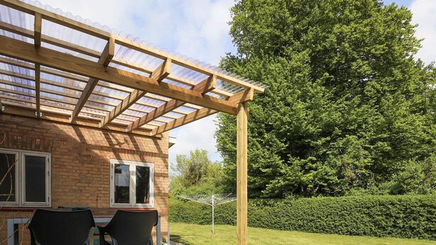 Een houten pergola met een glazen dak boven een terras, met twee stoelen en een tafel.