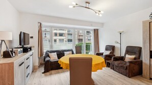 Una sala de estar moderna con una mesa redonda, un sofá y dos sillones. Hay un televisor sobre un mueble de madera, una lámpara de pie y una ventana con cortinas.