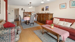 A living room with a red sofa, patterned armchair, and a wooden coffee table. There is a dining area with a table and chairs, and a kitchen area with cabinets and a microwave. - Flat With Terrace & Play Area (Blankenberge)