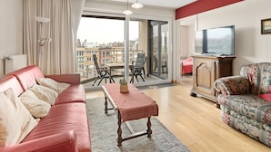 A modern living room with a red sofa, a patterned armchair, a wooden coffee table, and a television on a wooden cabinet. - Flat With Terrace & Play Area (Blankenberge)