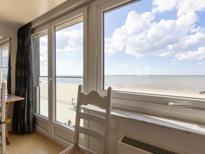 A room with a large window overlooking a beach, a chair, and a small table. - Nice Apartment With Shared Pool (Middelkerke)
