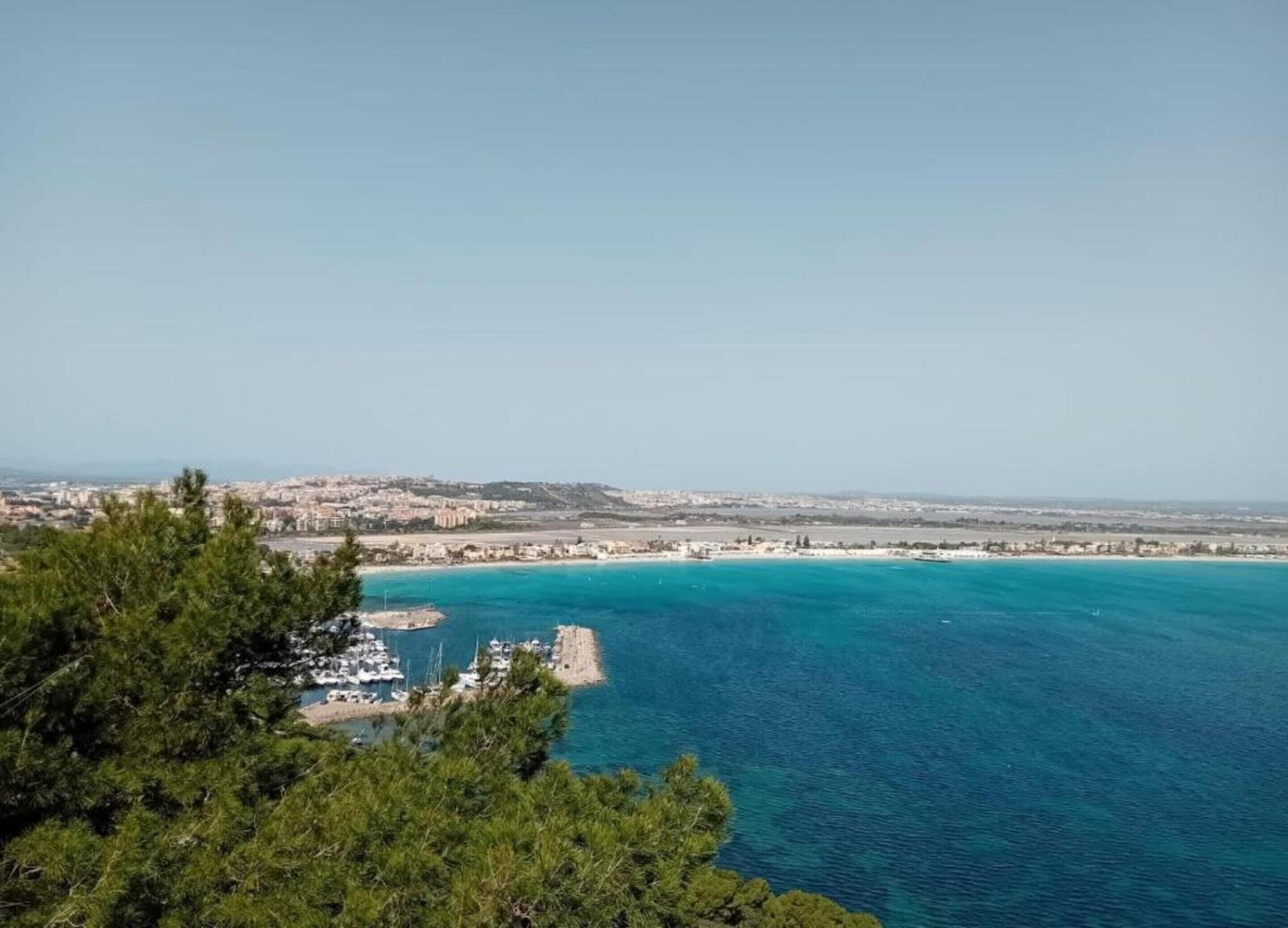 A coastal view with a marina, clear blue waters, and a distant cityscape.