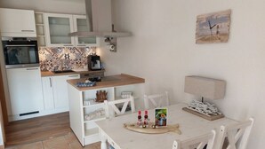 A compact kitchen with white cabinets, a coffee machine, and a dining area with a table and chairs. - Sky Blue Comfortable Holiday Residence (Friedrichskoog)