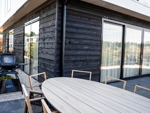 A modern outdoor patio with a wooden table and chairs, a grill, and a glass sliding door leading to a balcony.