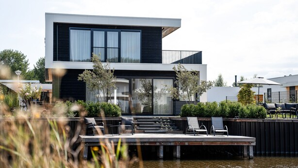 Una casa moderna de dos pisos con un muelle que se adentra en el agua, rodeada de vegetación.