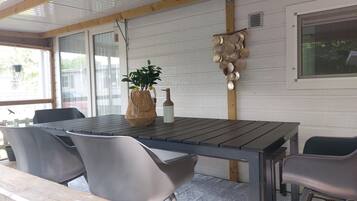 Een overdekt terras met een tafel, stoelen, een plant in pot en een decoratief object aan de muur.