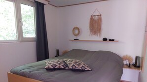 Une chambre à coucher avec un lit, une fenêtre avec des rideaux, une étagère murale décorée de divers objets, et une table de chevet avec une lampe.