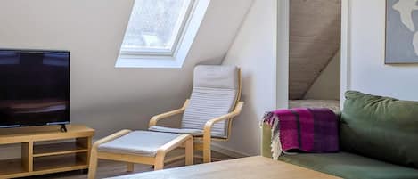A room with a TV, a wooden table, a cushioned chair, a sofa, and a window.