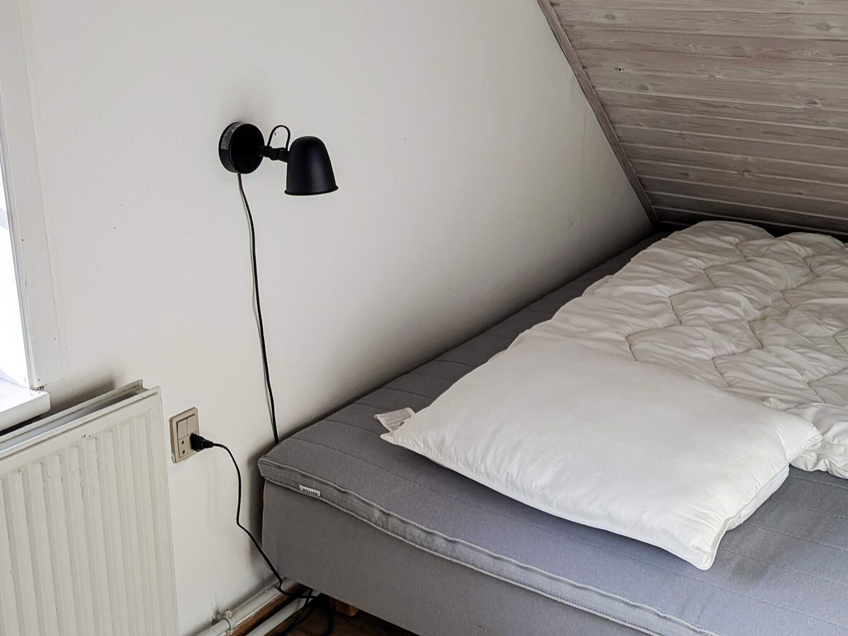 A small, single-bed room with a sloped ceiling, a white radiator, and a black wall-mounted lamp.