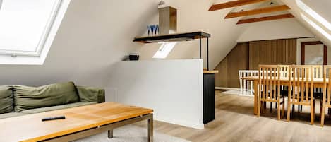 An attic room with a sloped ceiling, a dining table with chairs, a sofa, and a window.