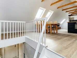 Avoin, loft-tyylinen huone, jossa on puulattia, valkoinen kaide ja ruokailutila, jossa on puutuoleja ja pöytä.