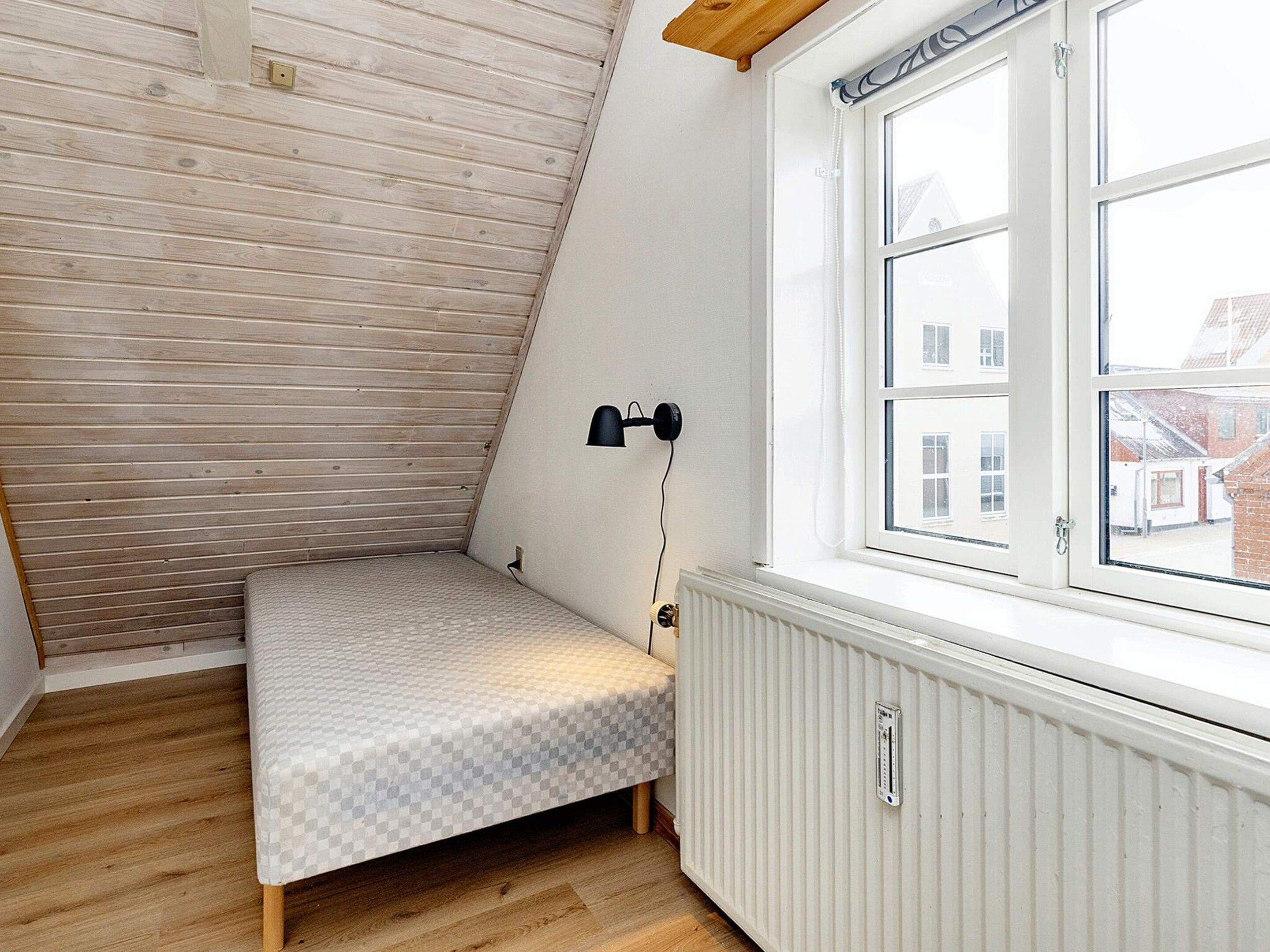 Et lille, skråt værelse med en enkelt seng, en radiator og et vindue med udsigt til bygninger.