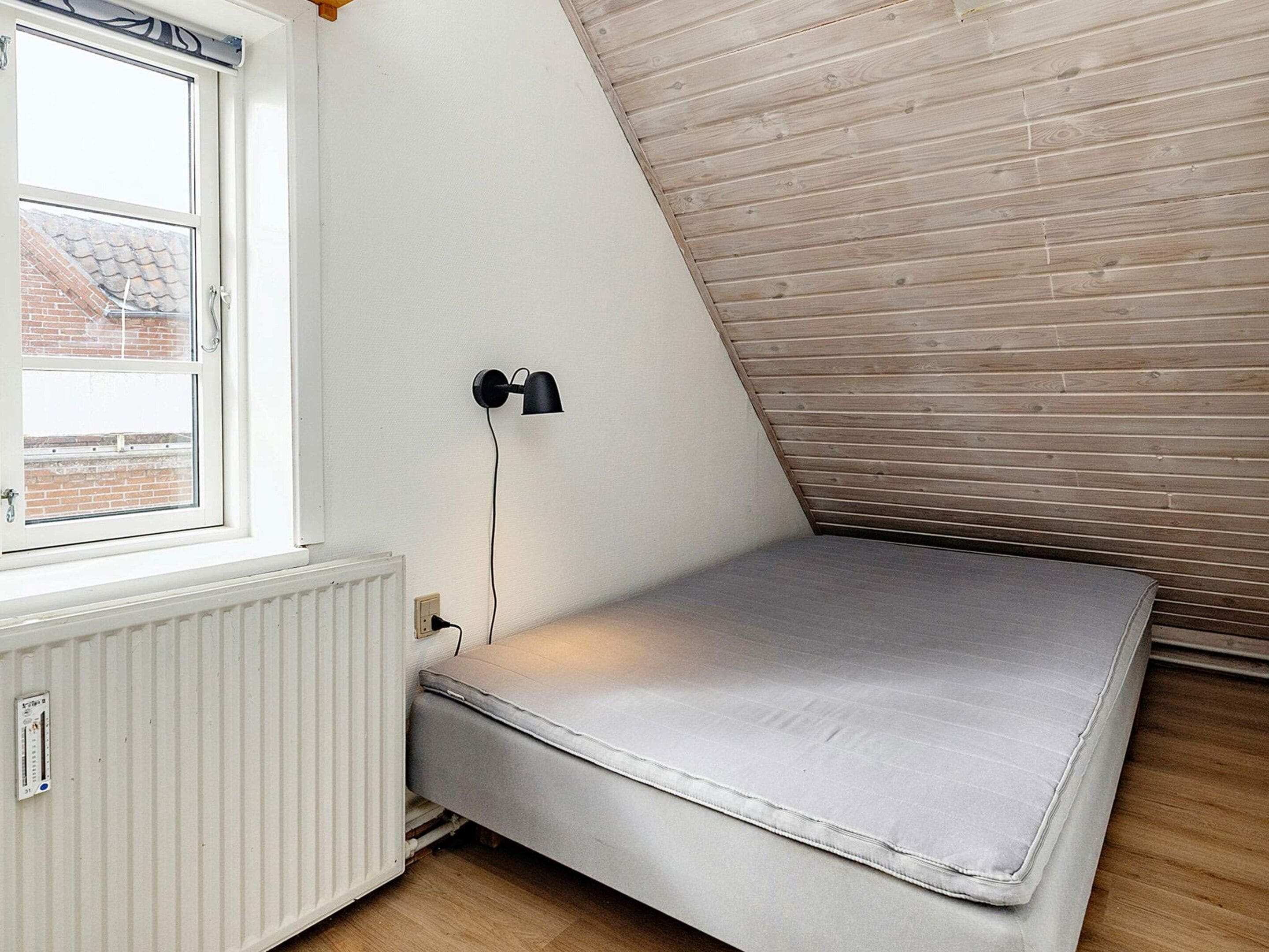 Et værelse med skråvæg, et vindue med udsigt til en murstensbygning, en vægmonteret lampe og en madras.
