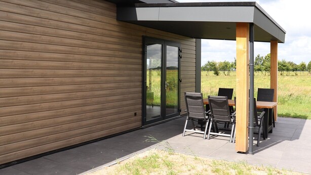 Una moderna veranda esterna con tavolo e sedie in legno, una porta vetrata che dà su un prato erboso e un’area relax coperta con vista sul paesaggio.