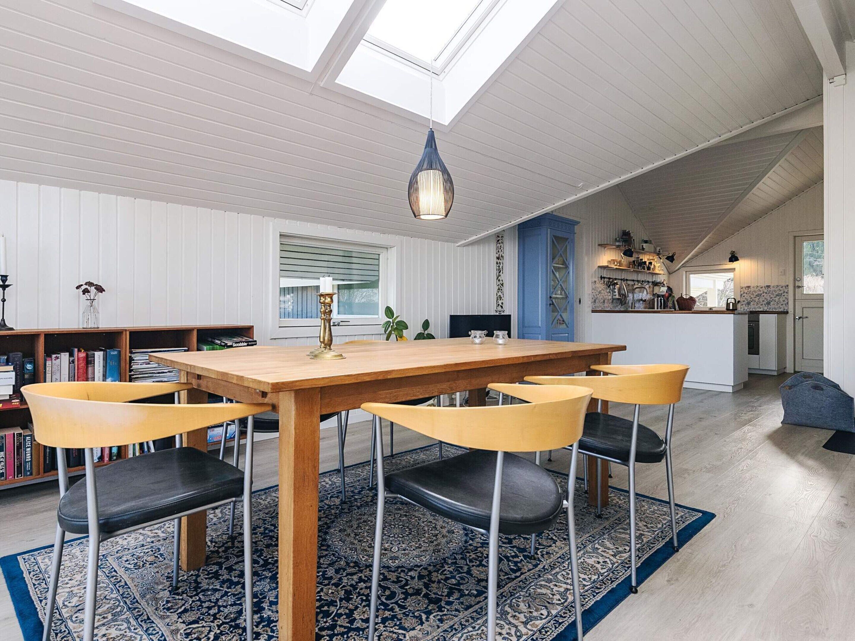Uma sala de jantar com mesa e cadeiras de madeira, um tapete azul estampado e um claraboia acima.